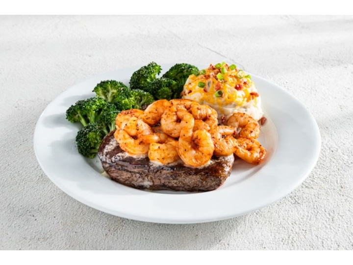 A bunch of grilled shrimp on top of a piece of steak with broccoli and a baked potato on a white plate.