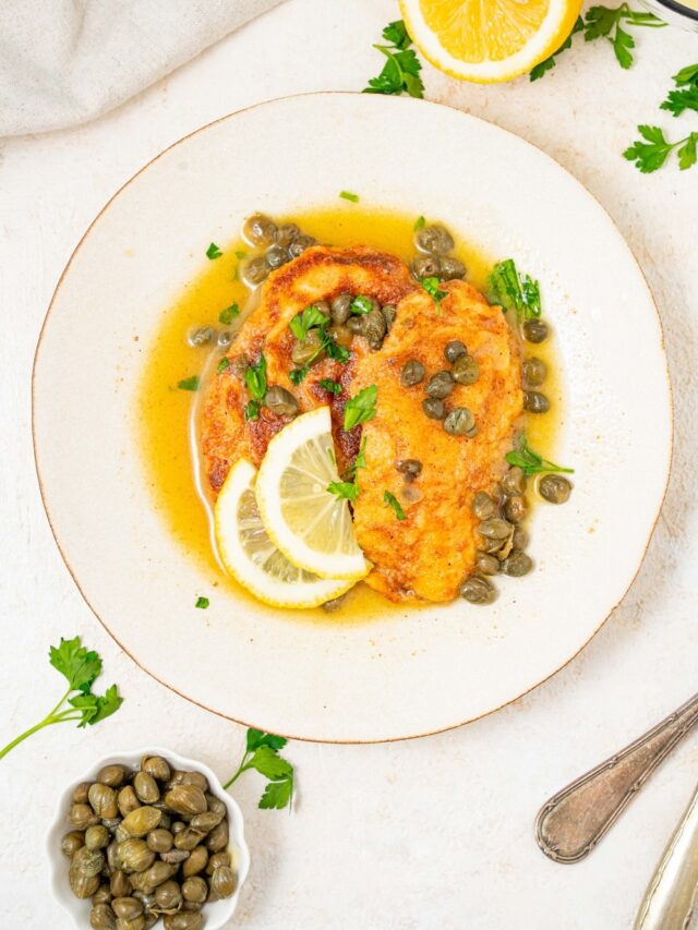 An overhead shot of a plate of two fried calamari steaks with a lemon caper sauce. The plate is garnished with sliced lemon and fresh parsley. There is a bowl of capers next to the plate.