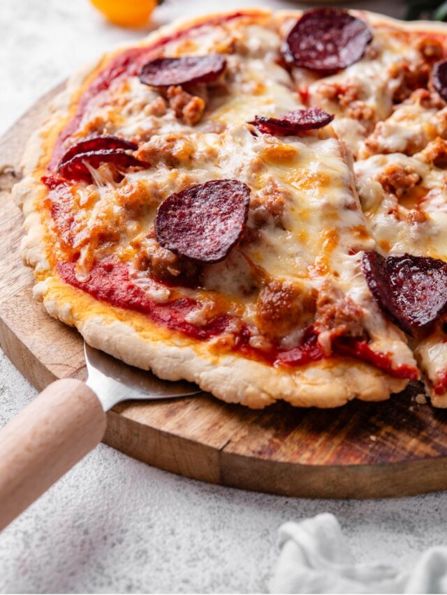 A whole protein pizza topped with cheese, sausage, and pepperoni on a wooden server. There is a pizza server inserted under a slice of pizza.