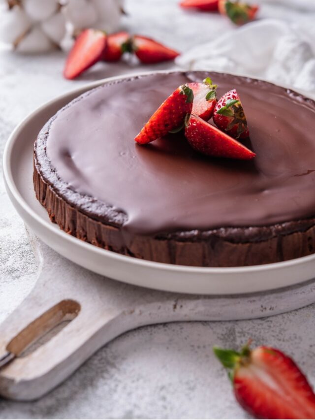 A whole chocolate protein cake topped with sliced strawberries. The cake is served on a white ceramic plate that sits on a white wooden serving board.