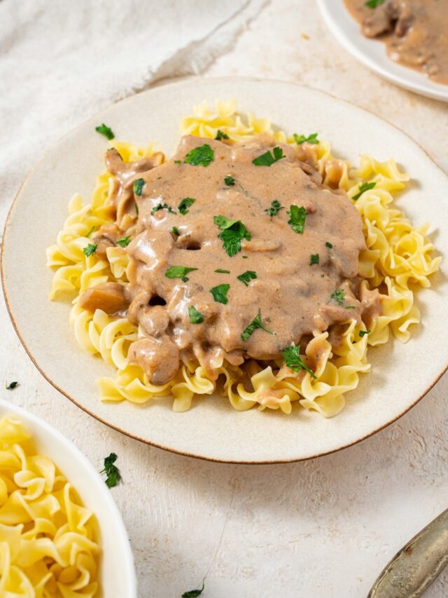 A plate filled with crock pot cubed steak and gravy on a bed of egg noodles with fresh parsley on top. There is an additional plate of egg noodles and an additional plate of cube steak in gravy on either side.