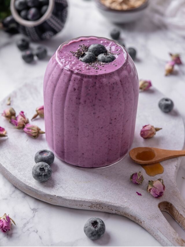 A ribbed glass filled with a high fiber smoothie served on a wooden board. The smoothie is topped with blueberries and chia seeds and there are blueberries and dried flower buds scattered around the board.