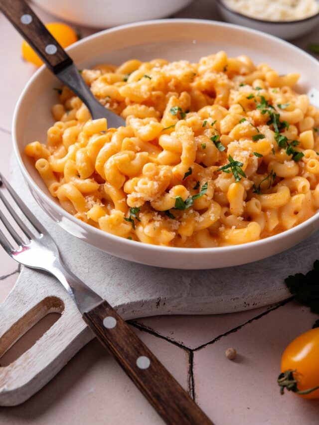 A bowl of protein mac and cheese on a white wooden server. There is a fork inserted into the macaroni and another fork next to the bowl. There is a second bowl of mac and cheese in the background.