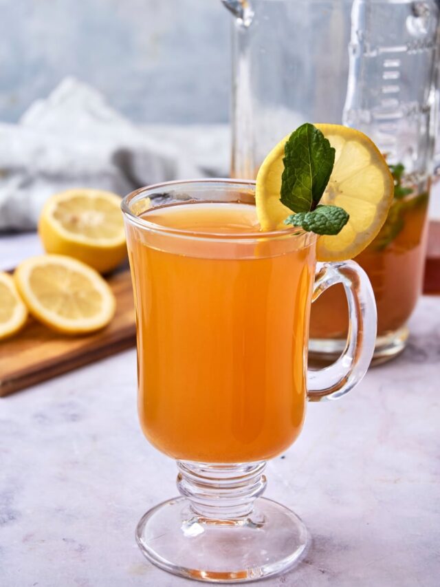 A glass tea mug filled with flu bomb tea garnished with fresh mint and a slice of lemon. There is a glass pitcher of flu bomb tea and a wooden board with sliced lemon in the background.