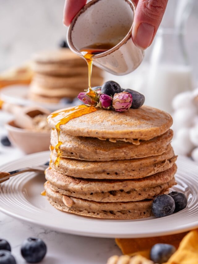 A stack of five high fiber pancakes on a white ceramic plate. The pancakes are topped with blueberries and dried flower buds and there is a hand pouring maple syrup from a ceramic ramekin over the pancakes.