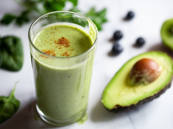 A green smoothie in a glass cup with an avocado next to it.