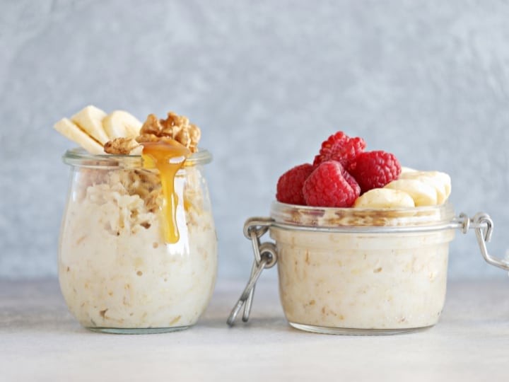 Two cups of overnight oats, one has banana and peanut butter on top and the other has strawberries on top.