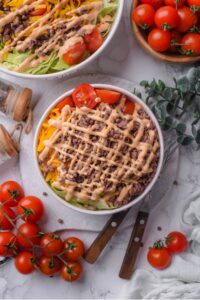 An overhead shot of a bowl of Big Mac salad filled with shredded lettuce, shredded cheddar cheese, cherry tomatoes, and ground beef drizzled with Big Mac salad dressing. There are cherry tomatoes on the vine next to the bowl, with another large bowl of Big Mac salad next to it.