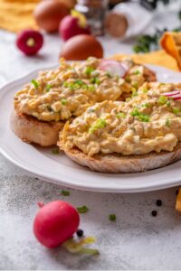 A plate of two slices of toasted bread with healthy egg salad on top. The egg salad is garnished with green onion and sliced radishes, with whole radishes surrounding the plate.