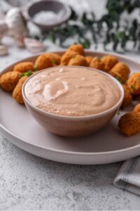 A small ceramic bowl of low calorie big mac sauce served on a white plate with breaded popcorn chicken.