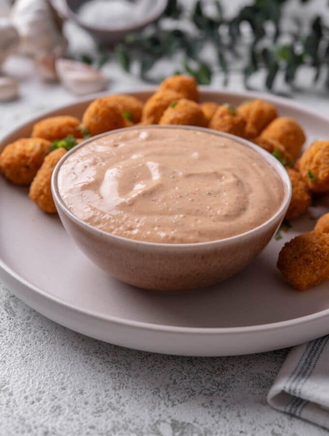A small ceramic bowl of low calorie big mac sauce served on a white plate with breaded popcorn chicken.