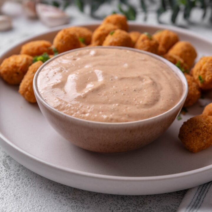 A small ceramic bowl of low calorie big mac sauce served on a white plate with breaded popcorn chicken.