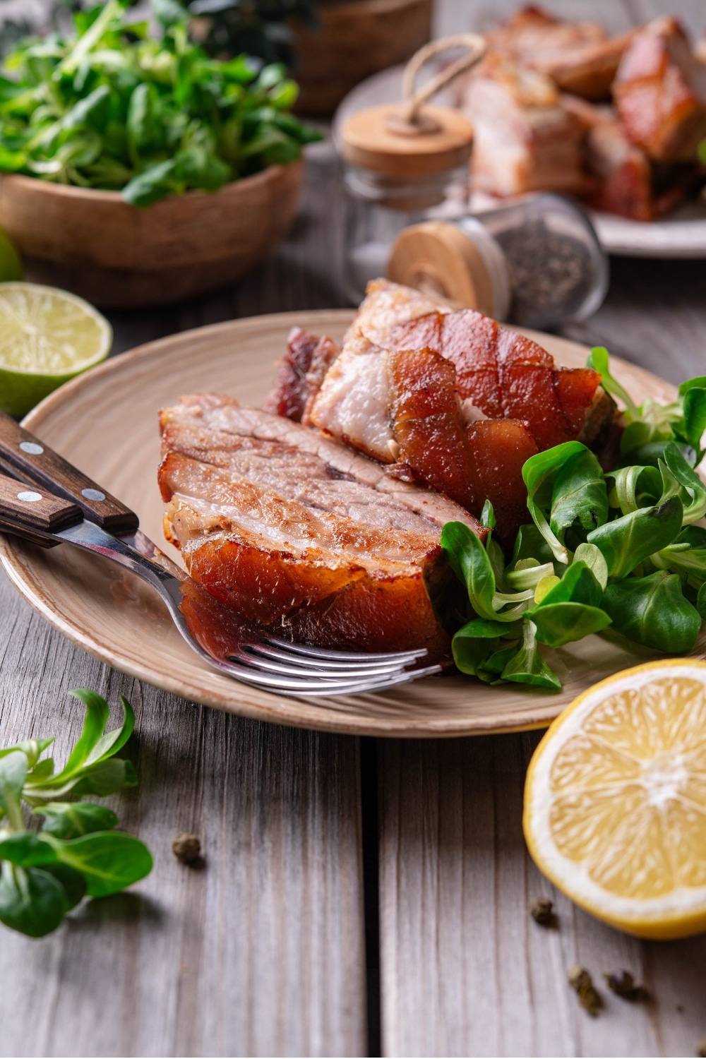 A tan ceramic plate with two pieces of sliced pork belly with a small side of greens. There are two forks on the plate and a halved lemon in front of the plate.