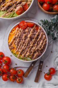 An overhead shot of a bowl of Big Mac salad filled with shredded lettuce, shredded cheddar cheese, cherry tomatoes, and ground beef drizzled with Big Mac salad dressing. There are cherry tomatoes on the vine next to the bowl, with another large bowl of Big Mac salad next to it.