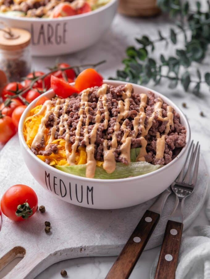 A bowl of Big Mac salad with shredded lettuce, shredded cheddar cheese, and ground beef drizzled with Big Mac salad dressing on top. The bowl has the word "Medium" written on it, and is served on a ceramic platter. There is another Big Mac salad bowl in the background with the word "large" written on it.