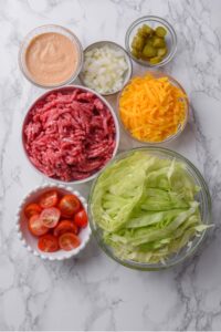 An overhead shot of several bowls of various sizes and shapes containing the ingredients for Big Mac salad including raw beef, shredded iceberg lettuce, shredded cheddar cheese, diced white onion, halved cherry tomatoes, chopped pickles, and Big Mac salad dressing.
