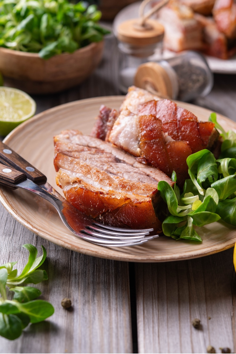 A tan ceramic plate with two pieces of sliced pork belly with a small side of greens. There are two forks on the plate and a halved lemon in front of the plate.