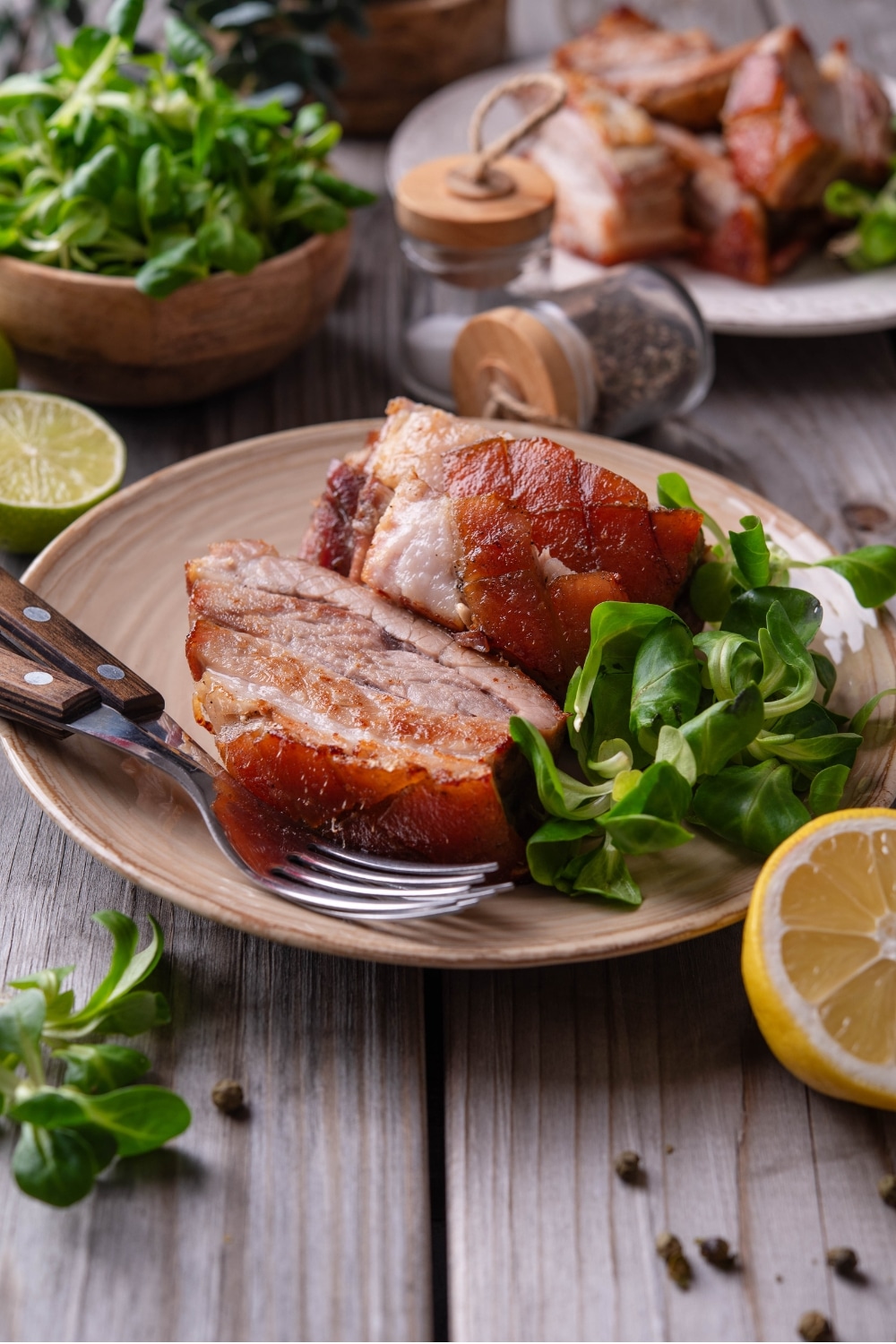 A tan ceramic plate with two pieces of sliced pork belly with a small side of greens. There are two forks on the plate and a halved lemon in front of the plate.