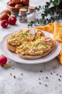 A plate of two slices of toasted bread with healthy egg salad on top. The egg salad is garnished with green onion and sliced radishes, with whole radishes surrounding the plate.