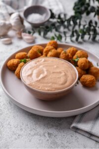 A small ceramic bowl of low calorie big mac sauce served on a white plate with breaded popcorn chicken.