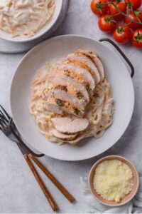 An overhead shot of a ceramic plate filled with spaghetti in low calorie alfredo sauce, topped with sliced grilled chicken. There are two forks and a small bowl of grated parmesan cheese next to the bowl.