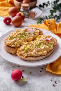 A plate of two slices of toasted bread with healthy egg salad on top. The egg salad is garnished with green onion and sliced radishes, with whole radishes surrounding the plate.