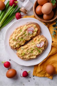 An overhead shot of a plate of two slices of toasted bread with healthy egg salad on top. The egg salad is garnished with green onion and sliced radishes, with whole radishes surrounding the plate. There is a small bowl of eggs next to the plate.