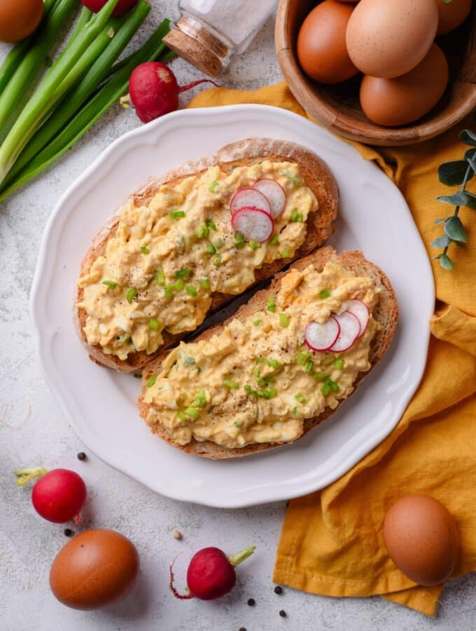 An overhead shot of a plate of two slices of toasted bread with healthy egg salad on top. The egg salad is garnished with green onion and sliced radishes, with whole radishes surrounding the plate. There is a small bowl of eggs next to the plate.