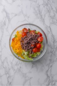 An overhead shot of a large glass bowl filled with shredded iceberg lettuce, halved cherry tomatoes, shredded cheddar cheese, and ground beef.