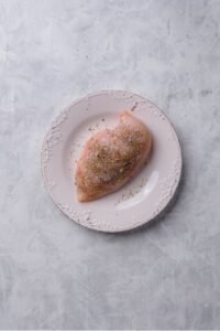 An overhead shot of a white plate with raw chicken breast seasoned with italian herbs, salt, and pepper
