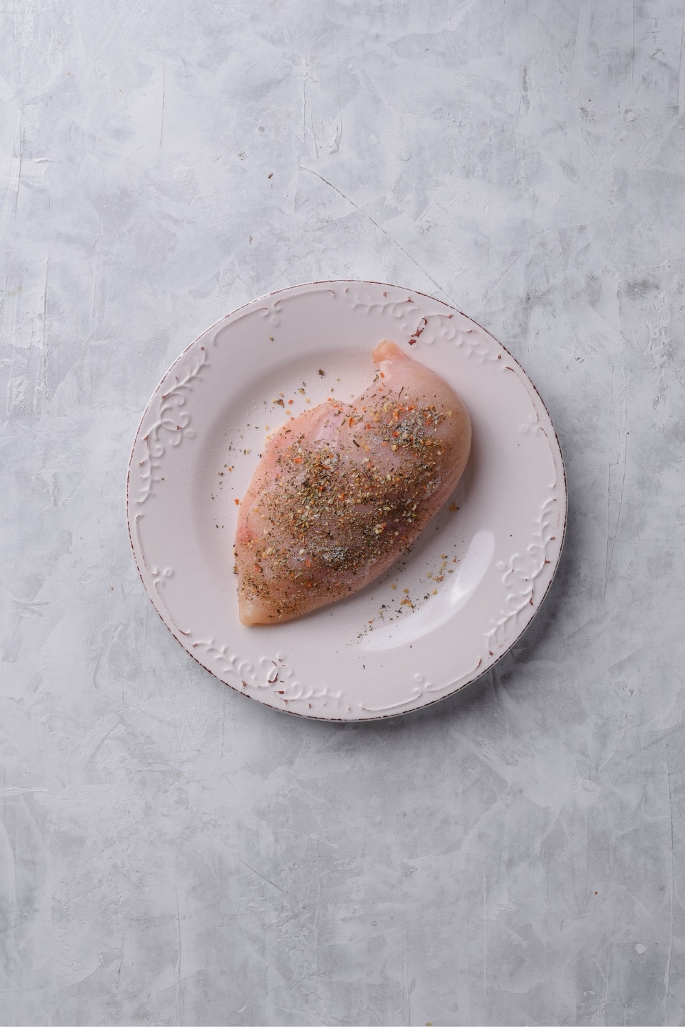 An overhead shot of a white plate with raw chicken breast seasoned with italian herbs, salt, and pepper