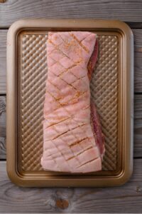 A scored pork belly topped with seasoning on a baking tray.