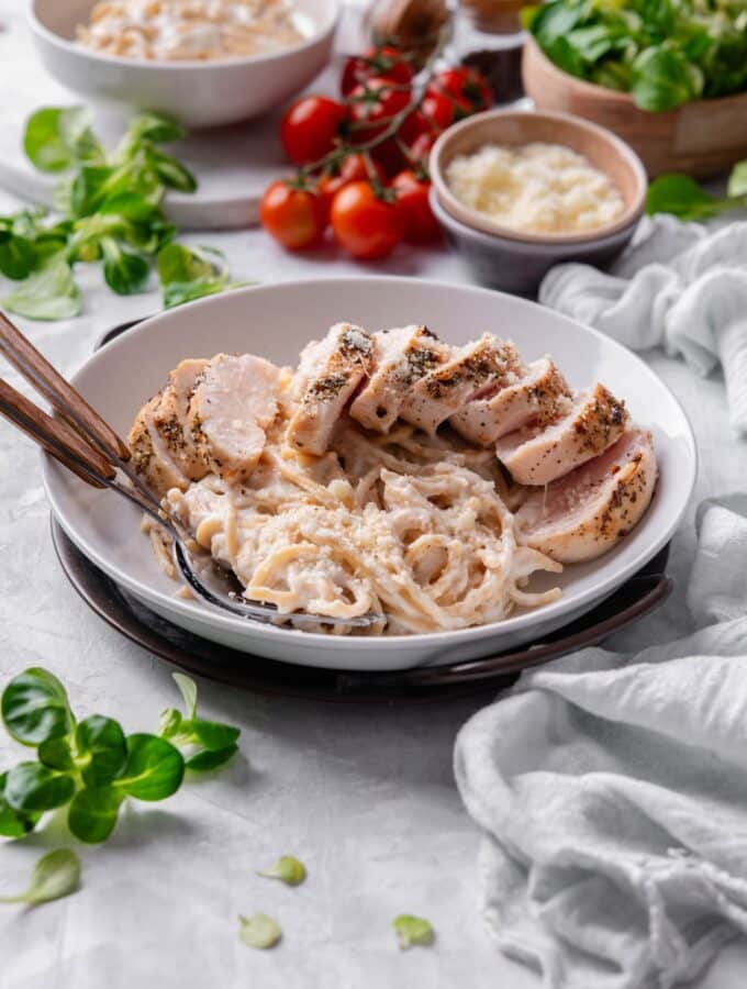 A ceramic plate filled with linguine in low calorie alfredo sauce, topped with sliced grilled chicken. There are two forks dipped into the pasta.