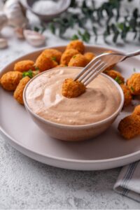 A small ceramic bowl filled with low calorie big mac sauce served on a plate with popcorn chicken. There is a fork dipping a piece of chicken into the sauce.