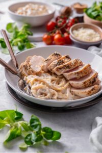 A ceramic plate filled with linguine in low calorie alfredo sauce, topped with sliced grilled chicken. There are two forks dipped into the pasta.