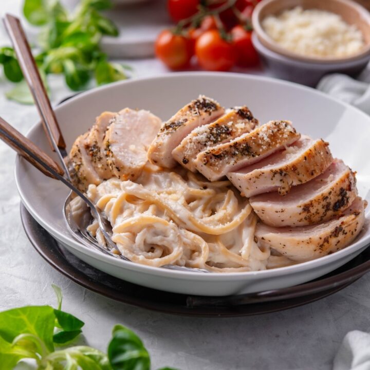 A ceramic plate filled with linguine in low calorie alfredo sauce, topped with sliced grilled chicken. There are two forks dipped into the pasta.