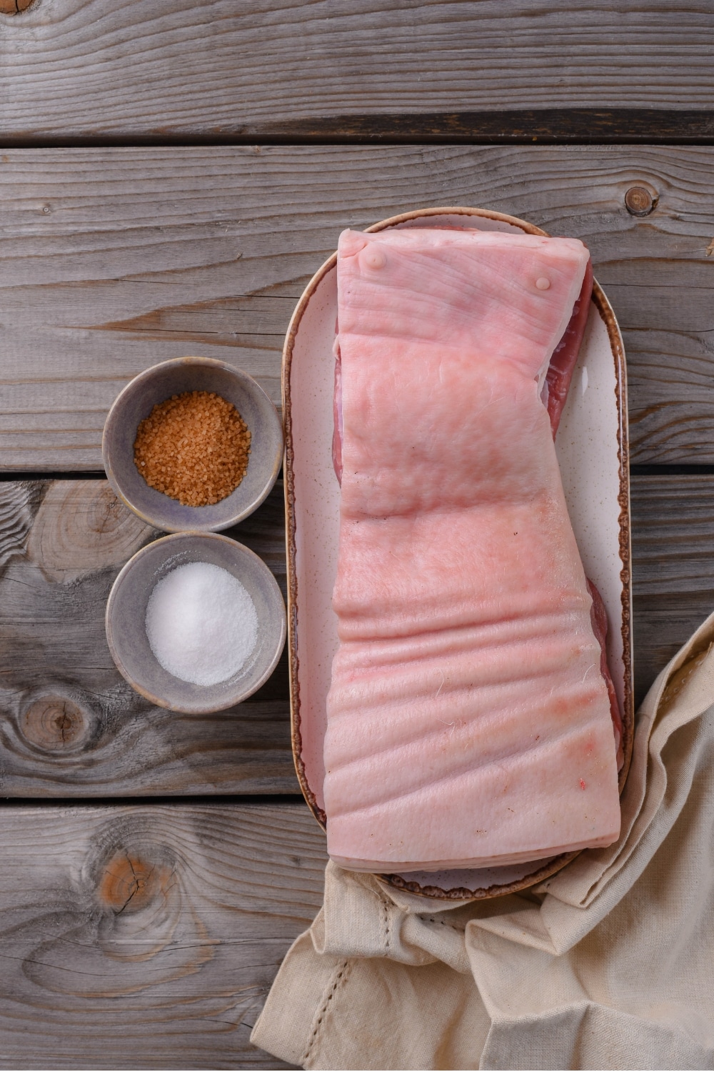 An overhead shot of a plate with raw pork belly, and two small bowls of brown sugar and salt.