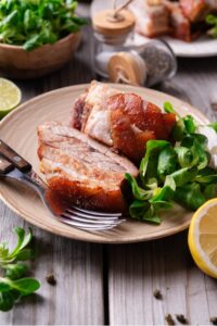 A tan ceramic plate with two pieces of sliced pork belly with a small side of greens. There are two forks on the plate and a halved lemon in front of the plate.