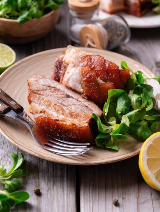 A tan ceramic plate with two pieces of sliced pork belly with a small side of greens. There are two forks on the plate and a halved lemon in front of the plate.