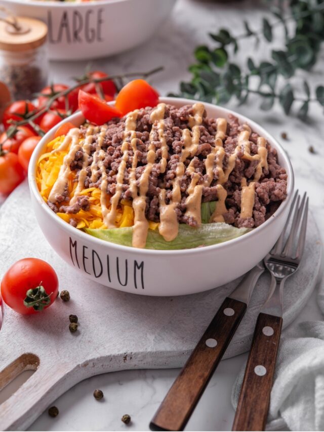 A bowl of Big Mac salad with shredded lettuce, shredded cheddar cheese, and ground beef drizzled with Big Mac salad dressing on top. The bowl has the word "Medium" written on it, and is served on a ceramic platter. There is another Big Mac salad bowl in the background with the word "large" written on it.