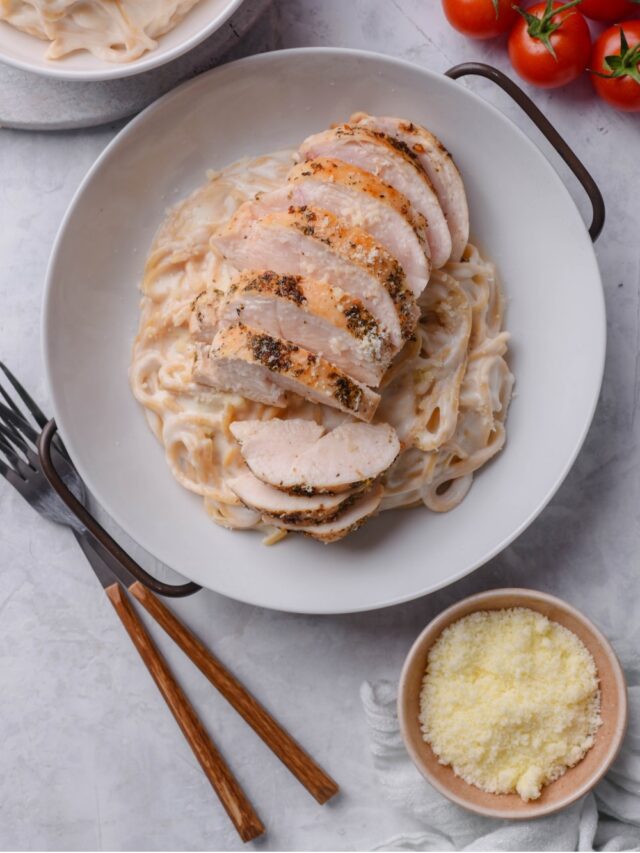 An overhead shot of a ceramic plate filled with spaghetti in low calorie alfredo sauce, topped with sliced grilled chicken. There are two forks and a small bowl of grated parmesan cheese next to the bowl.