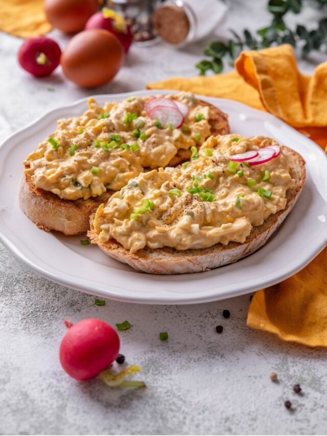 A plate of two slices of toasted bread with healthy egg salad on top. The egg salad is garnished with green onion and sliced radishes, with whole radishes surrounding the plate.