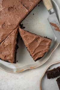 A chocolate cake topped with chocolate frosting that's been cut into slices using a knife that's on the plate. Some slices are missing.