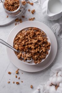 Overhead view of a bowl filled with granola and milk, and a spoon is in the bowl. The bowl is on a white plate and next to it is an open jar of granola.