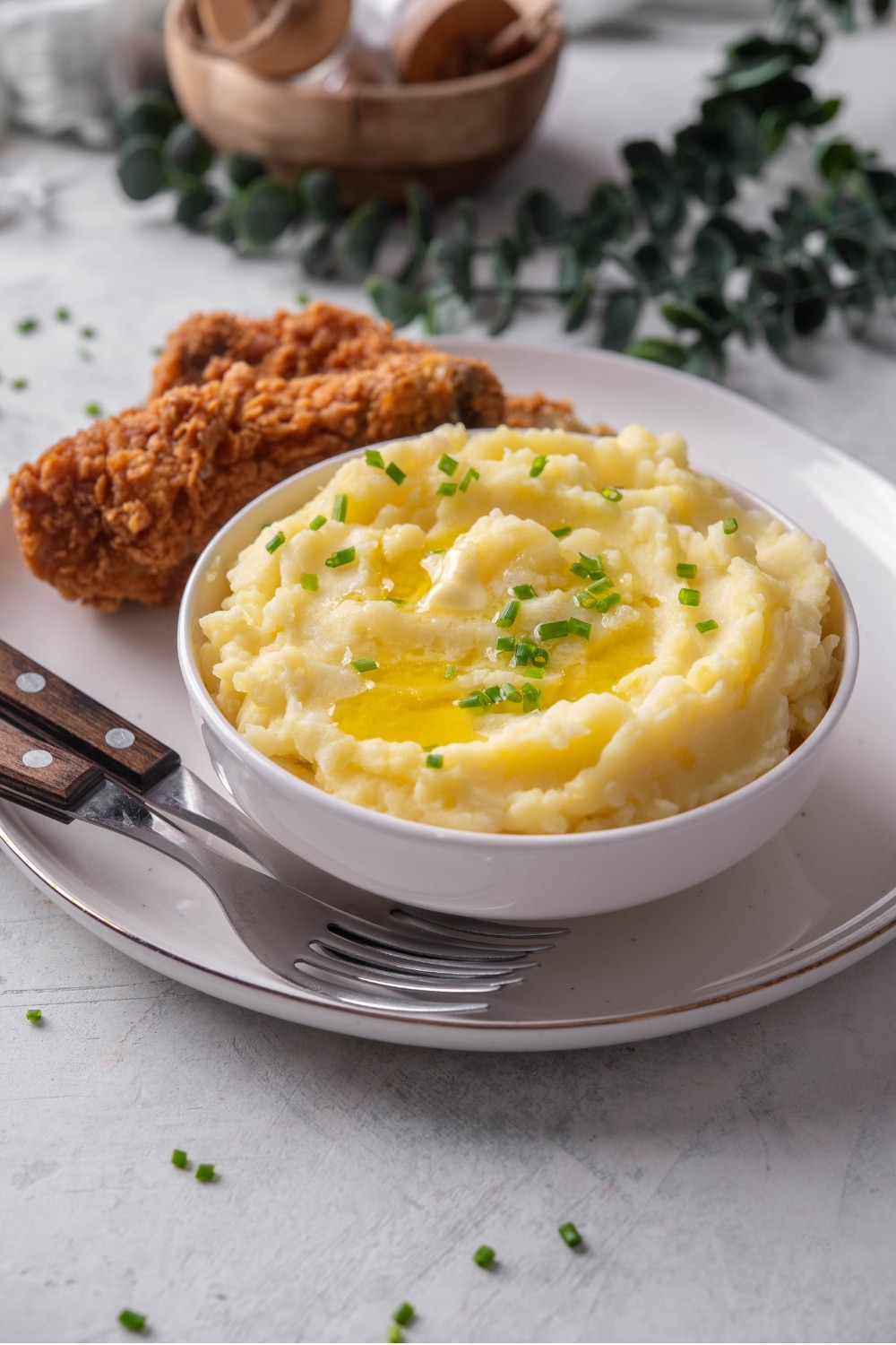 A white ceramic bowl filled with gold mashed potatoes with chives and melted butter on top. The bowl is on a ceramic plate with a piece of friend chicken next to the bowl. There are two forks tucked next to the bowl.