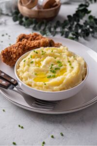 A white ceramic bowl filled with gold mashed potatoes with chives and melted butter on top. The bowl is on a ceramic plate with a piece of friend chicken next to the bowl. There are two forks tucked next to the bowl.