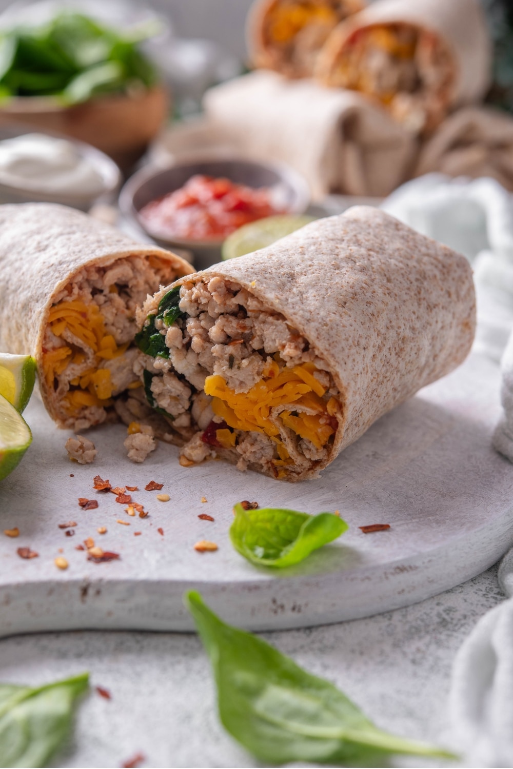 A high protein burrito sliced in half on a wooden server. The burrito has a whole wheat tortilla stuffed with ground turkey, cheese, egg whites, and spinach. There is spinach and red pepper flakes sprinkled on the server.