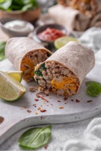 A high protein burrito sliced in half on a wooden server. The burrito has a whole wheat tortilla stuffed with ground turkey, cheese, egg whites, and spinach. There are lime wedges, spinach, and red pepper flakes sprinkled on the server.