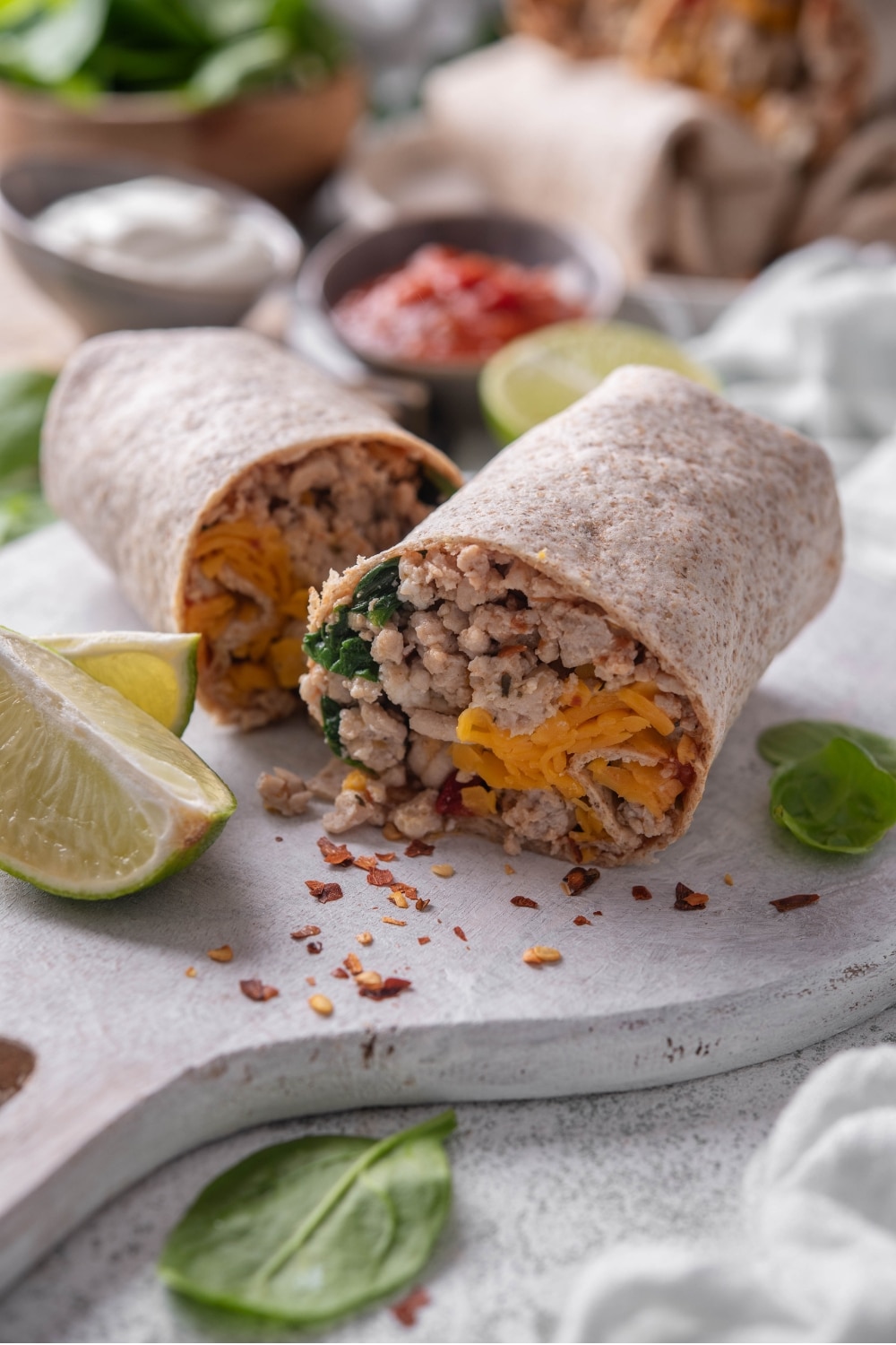 A high protein burrito sliced in half on a wooden server. The burrito has a whole wheat tortilla stuffed with ground turkey, cheese, egg whites, and spinach. There are lime wedges, spinach, and red pepper flakes sprinkled on the server.