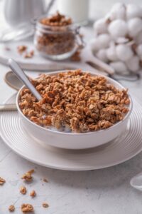 A white shallow bowl filled with granola and milk. There is a spoon in the bowl and the bowl is on a white plate.